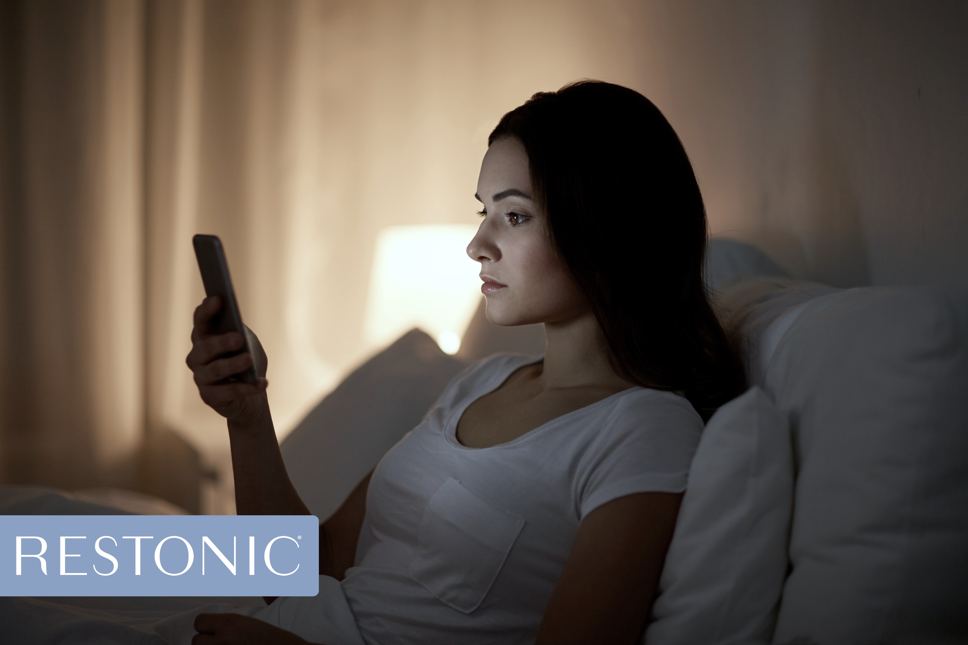 Woman sitting in bed looking at her mobile phone for new sleep hacks to help get through a sleep-deprived day.