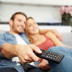 A husband and wife sitting on a couch watching television about how exercise will help them sleep better.