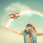 Girl watching a wooden airplane fly above her between a rainbow that stretches between her outstretched arms.