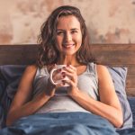 Beautiful young woman is holding a cup, looking at camera and smiling while lying in bed in the morning